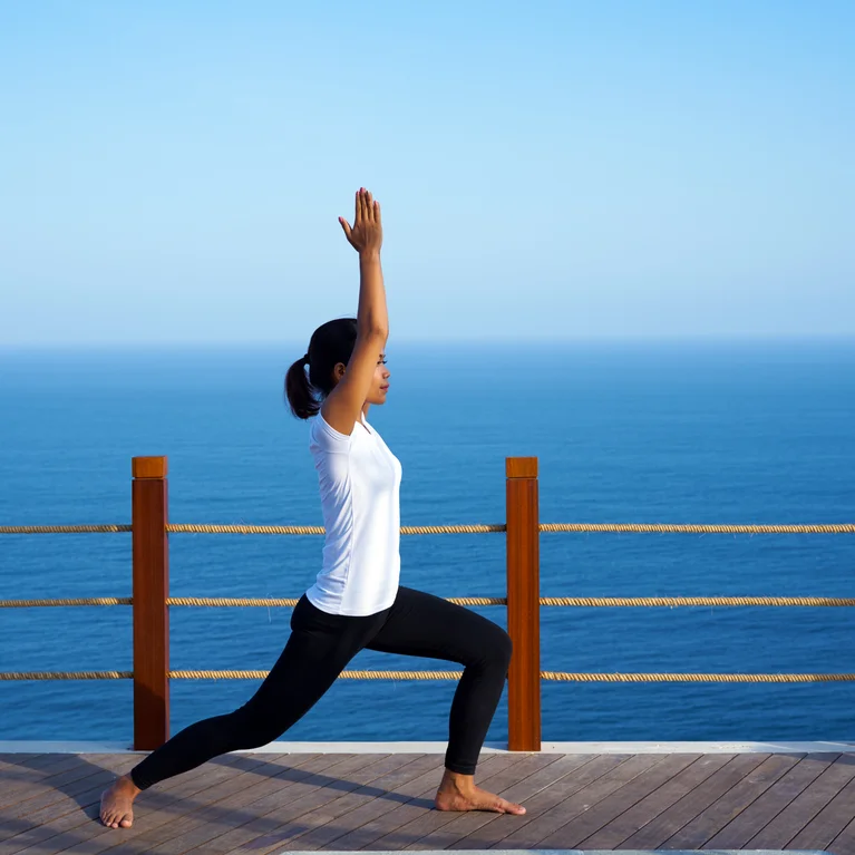 一个人在阳光明媚的木制露台上做瑜伽姿势,背景是蔚蓝的天空和轻柔的海浪。.