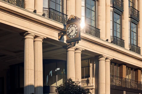 A classic clock on a beige building with tall columns and large windows. Natural sunlight highlights the architectural details and the clock’s face.