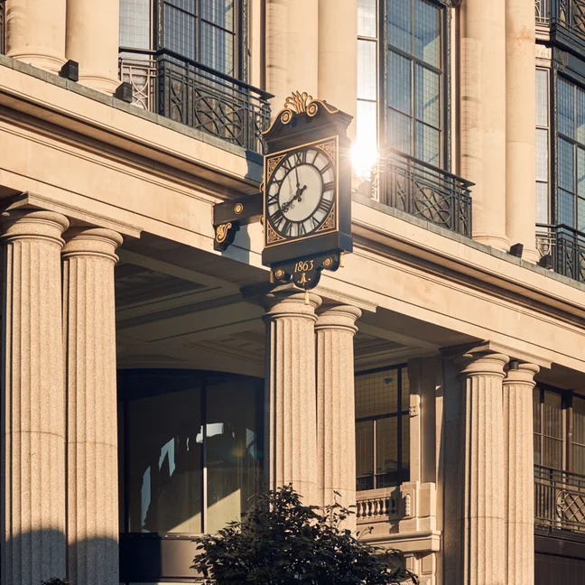 A classic clock on a beige building with tall columns and large windows. Natural sunlight highlights the architectural details and the clock’s face.