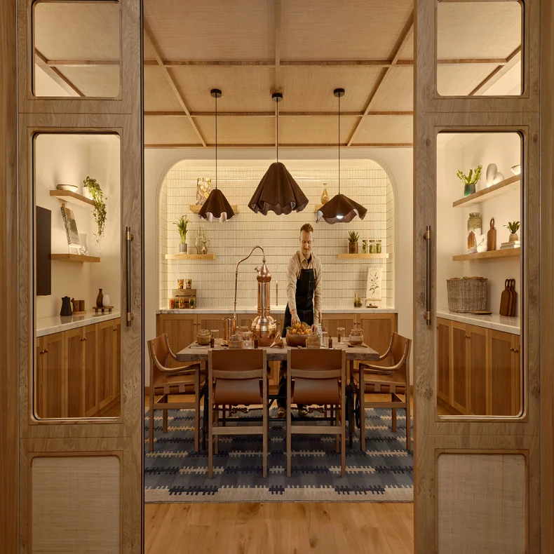 Person in an apron prepares a meal at a dining table in a modern kitchen with warm lighting, wooden cabinets, shelves, and black pendant lights.