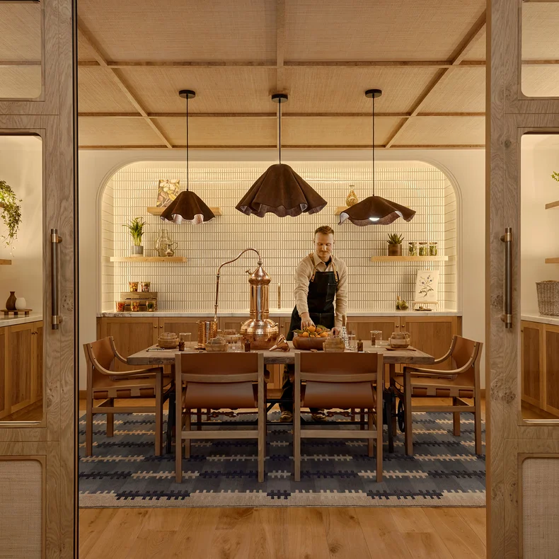 Person in an apron prepares a meal at a dining table in a modern kitchen with warm lighting, wooden cabinets, shelves, and black pendant lights.