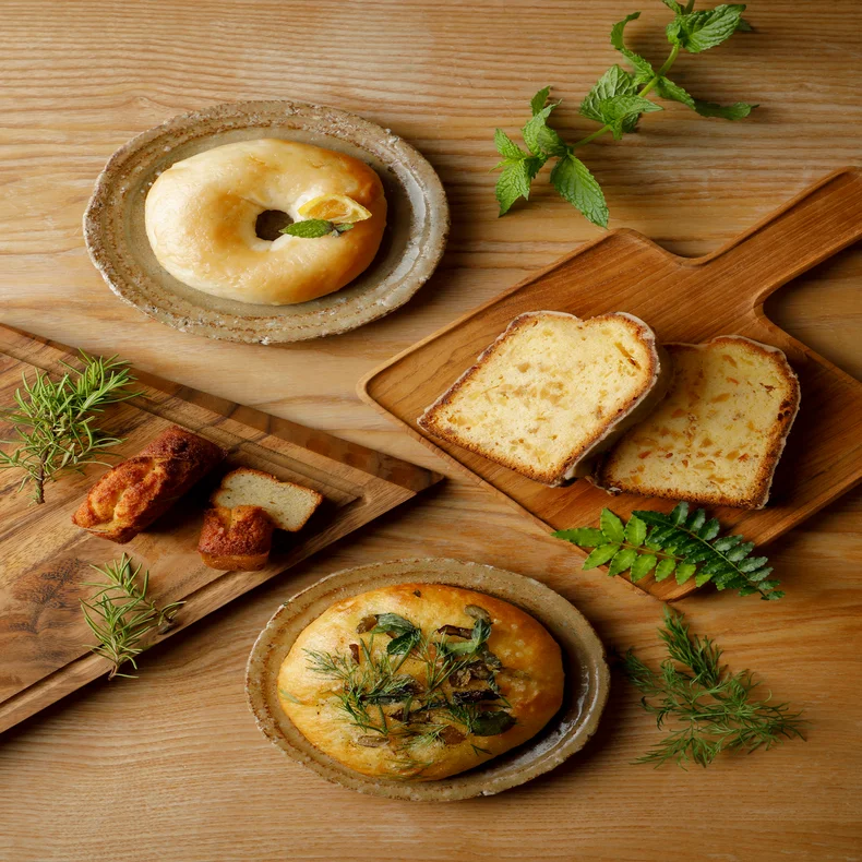 Selection of house-baked breads and patisserie on ceramic plates and wooden trays, finished with fresh herbs, arranged on a natural wood table for mindful enjoyment.