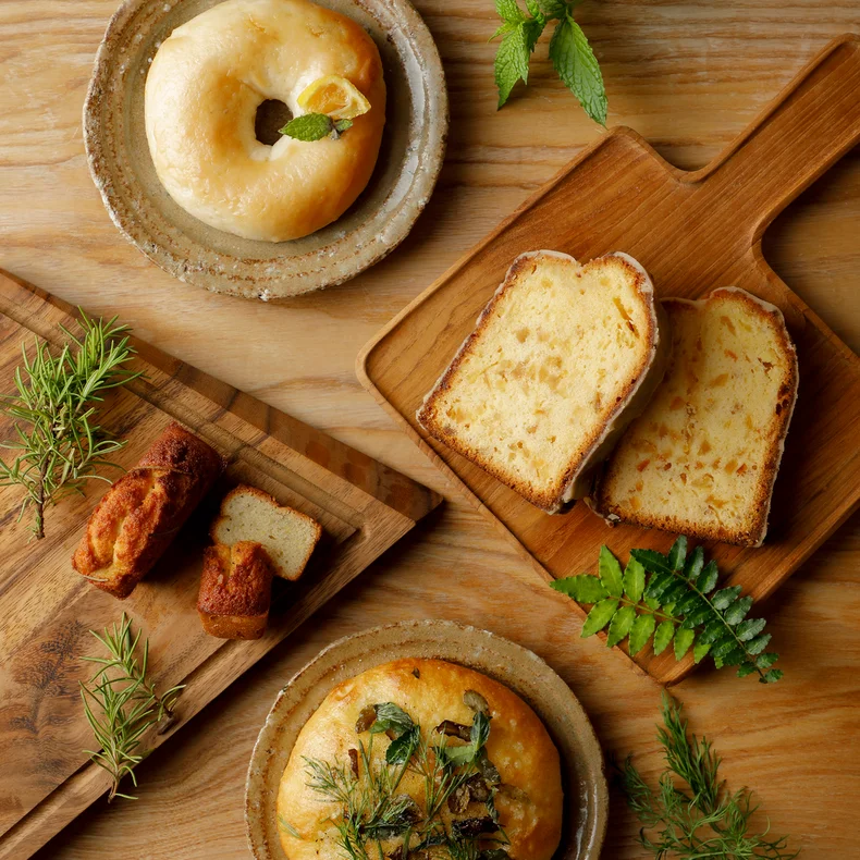 Selection of house-baked breads and patisserie on ceramic plates and wooden trays, finished with fresh herbs, arranged on a natural wood table for mindful enjoyment.