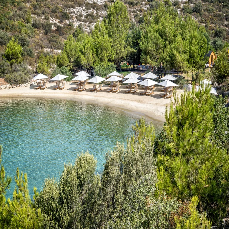 A tranquil beach with gentle waves, shaded umbrellas and sunbeds, framed by lush trees and natural stone hills. Accessible pathways lead to the shoreline.
