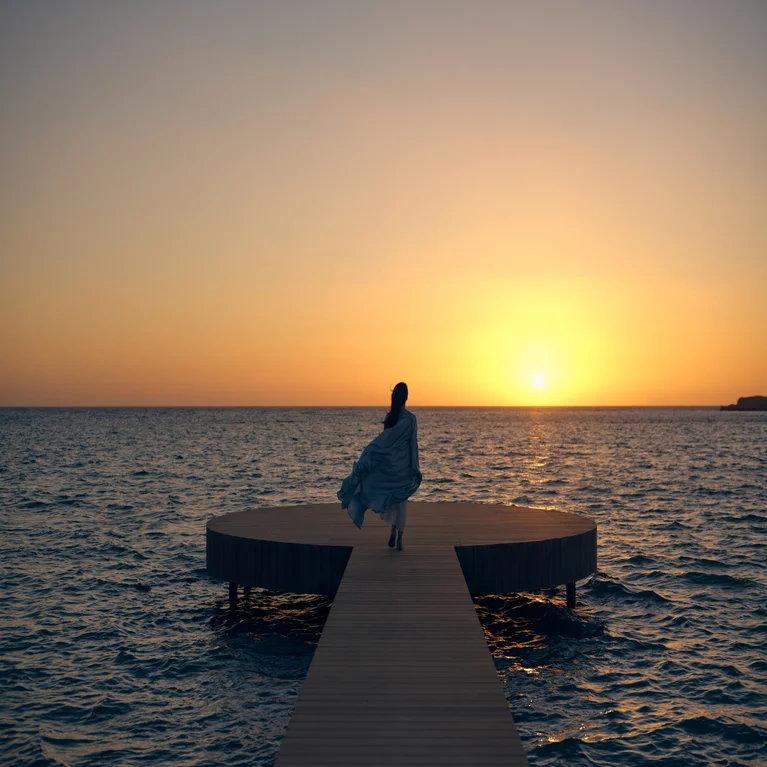 Guest enjoys a circular wooden pier overlooking the ocean at sunset, gentle waves surrounding the structure for a tranquil and natural setting.