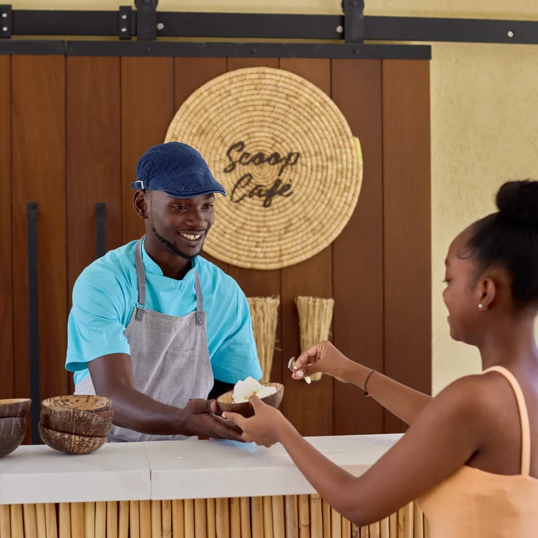 في مقهى سكوب كافيه، يقدم أحد الموظفين الآيس كريم لزبون عبر المنضدة؛ وبالقرب منه أوعية خشبية وموزع مياه، مع التركيز على الضيافة الفاخرة.