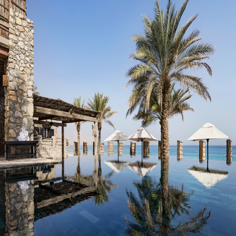 Infinity pool bordered by palm trees and white umbrellas, adjacent to a stone building overlooking calm sea under a blue sky. Accessible pathways connect poolside areas.