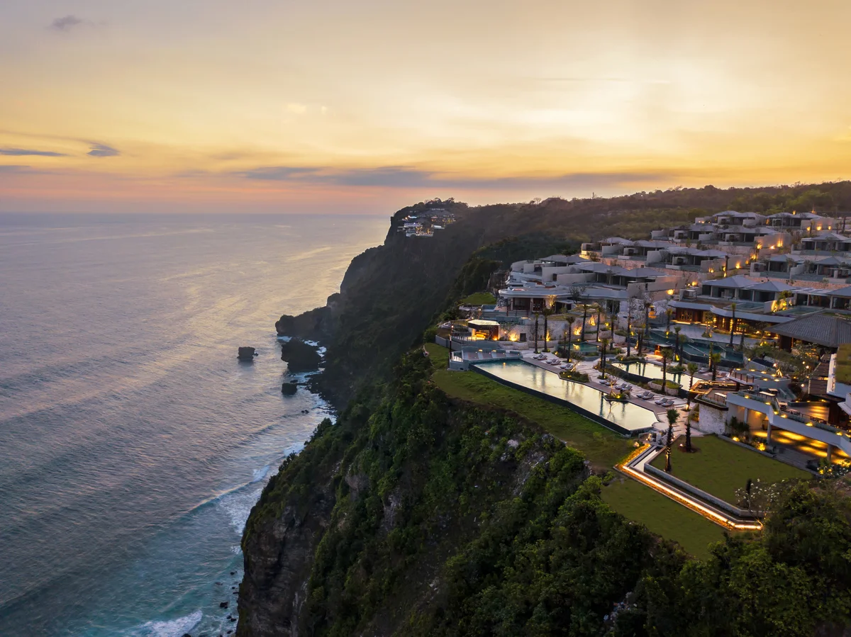 Villas stepping down the cliff at sunset, lit from within, with the pool at the edge and the sea below