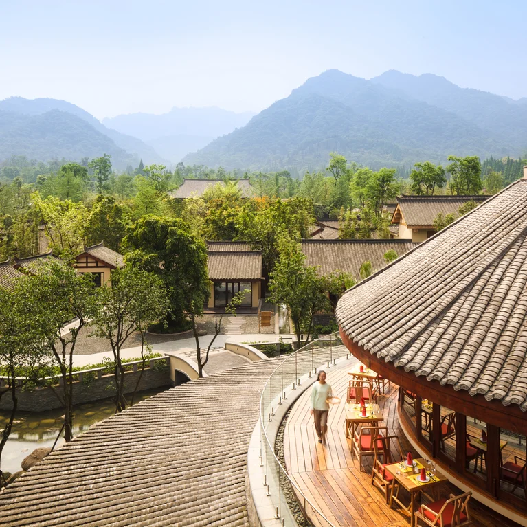 The rooftop terrace is furnished with tables and chairs and offers views of greenery, historic buildings and the misty mountains in the distance under clear skies....