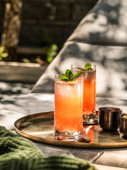 Glasses of pink-orange cocktail with ice and mint garnish are arranged on a tray in natural light, accompanied by metal cups and a folded green cloth.