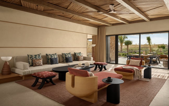 Living room with sofa, accent chairs, and coffee tables; patio doors lead to a terrace featuring outdoor seating and views of palm trees and green landscape.