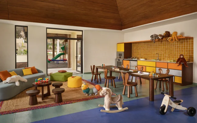 Children’s playroom featuring wooden toys, child-sized tables and chairs, a rocking horse, and play kitchen; large windows overlook a green outdoor playground.
