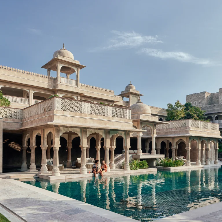 An elegant building with detailed arches and domes stands beside a calm outdoor pool, where two people are in the water beneath clear blue skies.