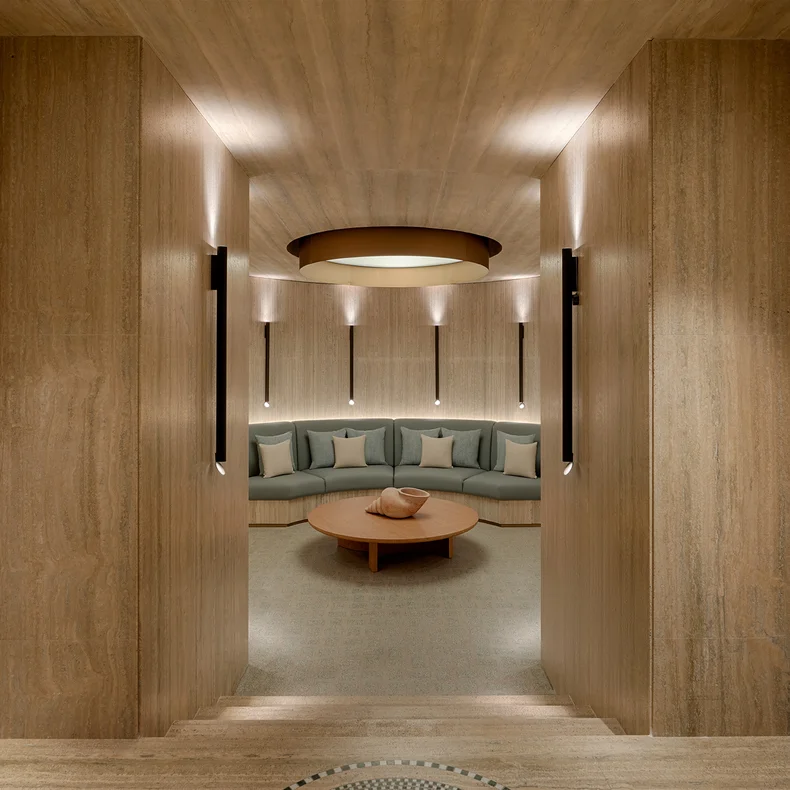 Lounge with timber walls, soft ambient light, a curved sofa and round coffee table at the center; designed for comfort and sustainable materials.