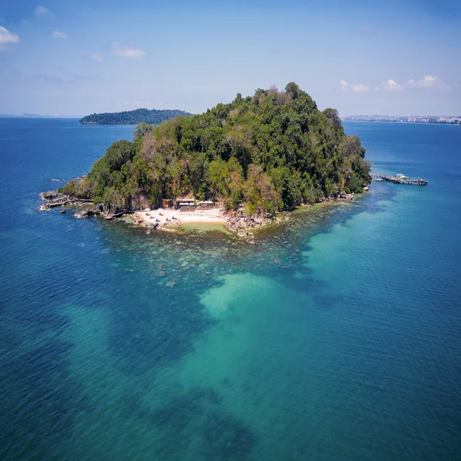 An island with green vegetation and a sandy shoreline, surrounded by clear blue sea, seen from above in bright sunlight.