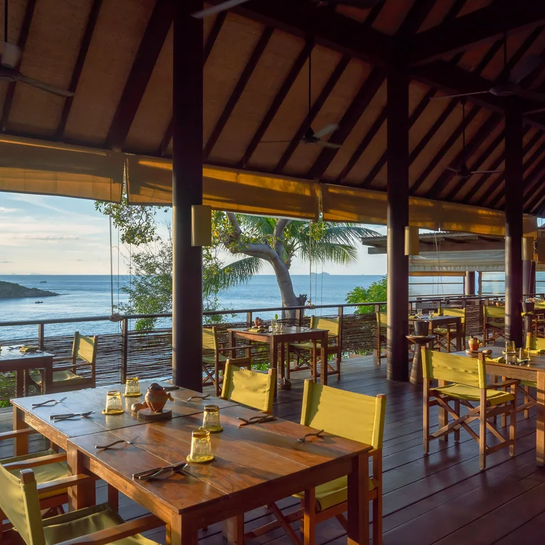 Restaurante con mesas y sillas de madera sostenible, vistas al océano, listo para cenar. La luz del sol se filtra en el interior, enmarcado por palmeras autóctonas en el exterior.