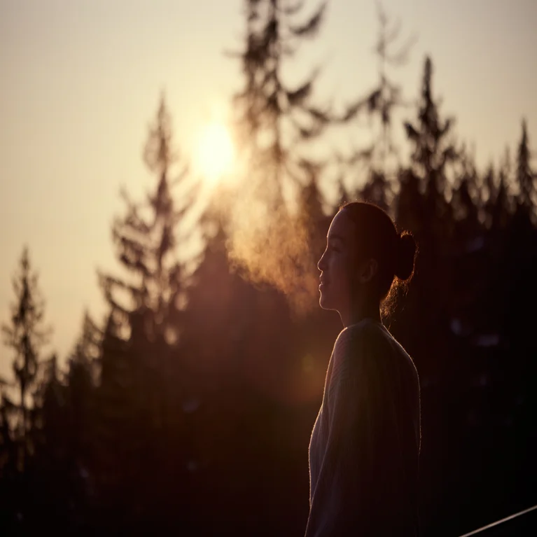 Someone enjoys fresh, cool air in a serene forest at dusk, with their breath visible and evergreen trees forming soft shapes in the fading light.