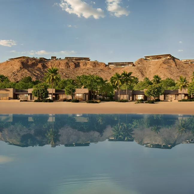 Villas and palm trees border a sandy shore. Still, clear water mirrors a nearby rocky hill and blue sky, evoking tranquility and connection to the natural surroundings.