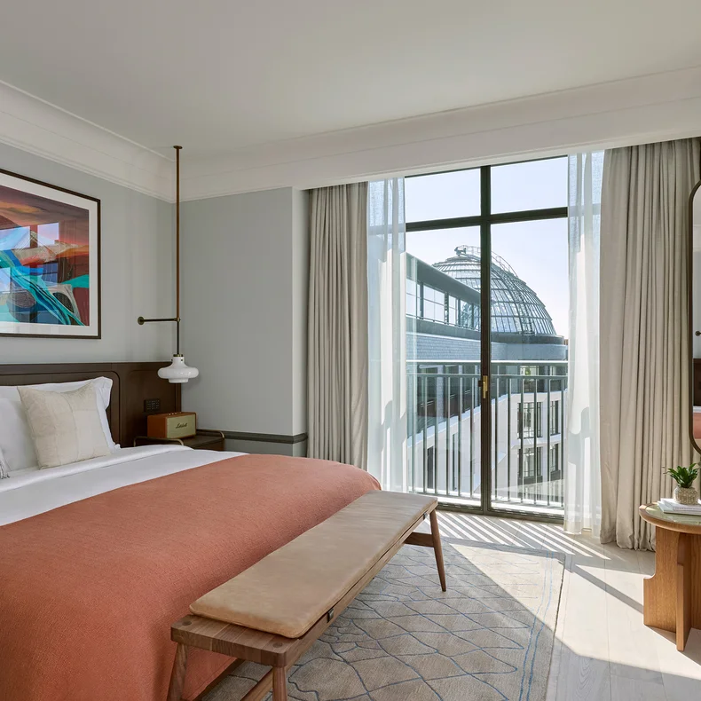King-sized bed with warm-toned blanket, natural wood details, twin mirrors, bedside tables and large window with sheer curtains framing a glass-domed view.