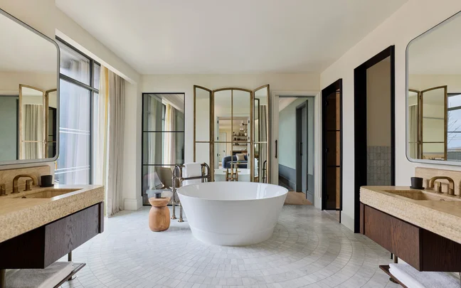 Bathroom with a central round soaking tub, dual vanities with mirrors, and glass doors offering access to an adjoining room; natural light highlights calm tones.