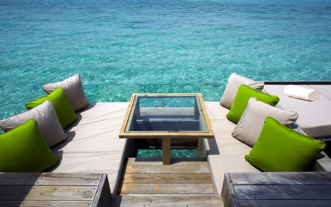 Seating with beige cushions and green pillows surrounds a glass table, set by the ocean with views of clear turquoise water. Accessible space for relaxation and connection.