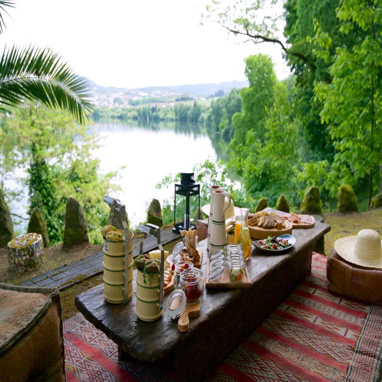 An outdoor picnic table set with fresh food and drinks sits by a calm river, surrounded by vibrant green trees and natural scenery.