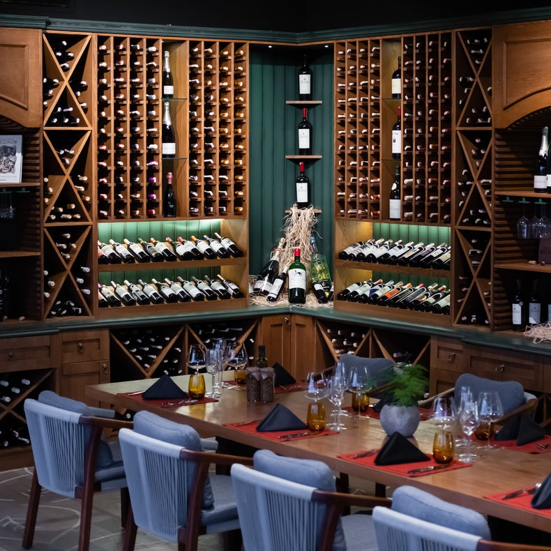 En la espaciosa bodega, las botellas de vino están ordenadas en estantes de madera, y frente a ellas hay una gran mesa con cubiertos, servilletas y asientos para los invitados.