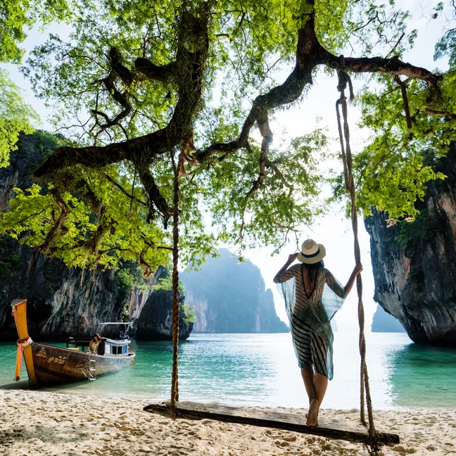 Un invité profite d'une balançoire ombragée sous un grand arbre, surplombant l'eau bleue claire et les falaises, avec un bateau en bois ancré sur la plage voisine.