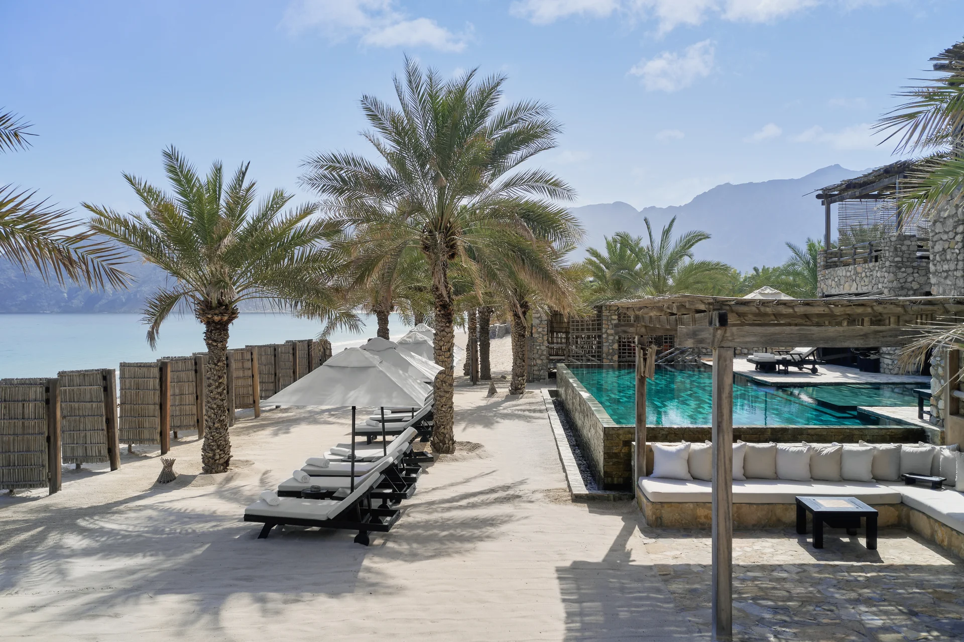 The Private Reserve from its sand terrace, date palms and canvas parasols shading a row of loungers beside a long green pool with a cushioned pergola daybed and the mountains beyond.