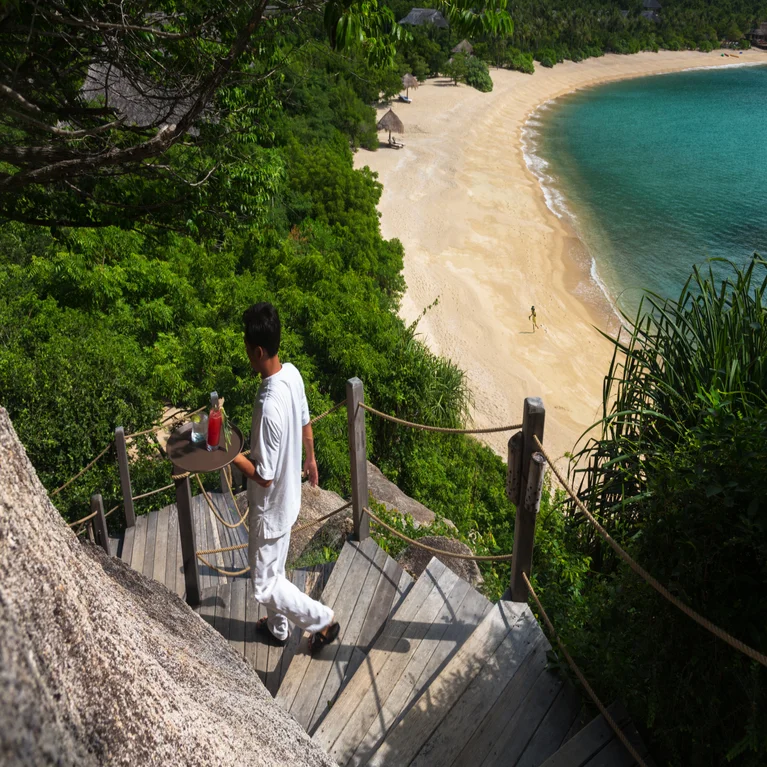 A team member brings refreshments down wooden steps toward a beach with sand and turquoise water, surrounded by abundant green foliage.
