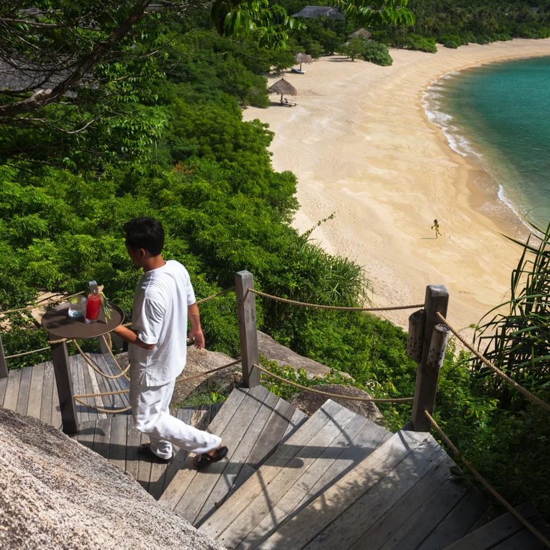 A team member brings refreshments down wooden steps toward a beach with sand and turquoise water, surrounded by abundant green foliage.
