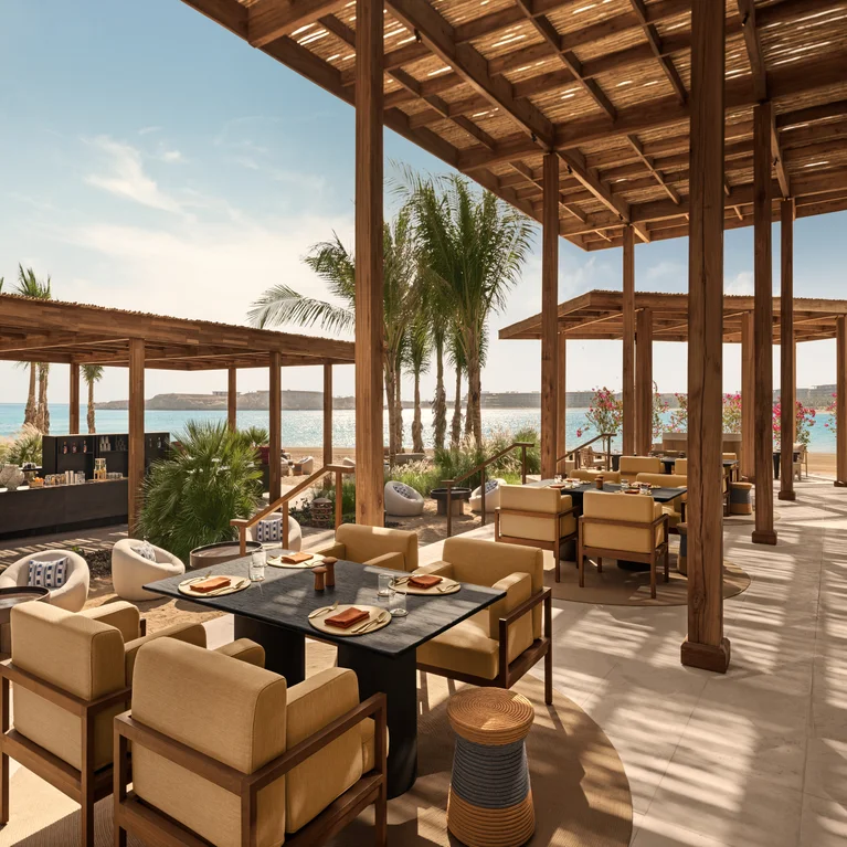 Dining area shaded by a wooden pergola features neutral-tone seating and dark tables, with palm trees and sea visible under clear skies, inviting relaxation.