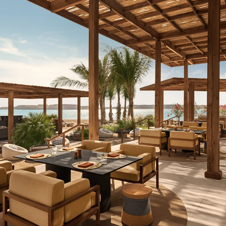 Dining area shaded by a wooden pergola features neutral-tone seating and dark tables, with palm trees and sea visible under clear skies, inviting relaxation.