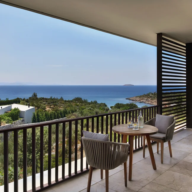 A serene terrace with a small round table and two chairs faces the sea, framed by lush plants, white buildings below, and an open blue sky.