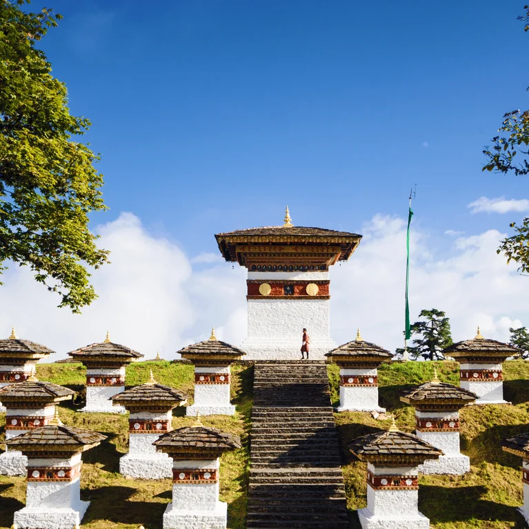 White chortens with gold roofs stand on a grassy hilltop beneath a blue sky, a central chorten with stair access inviting exploration in this serene landscape.