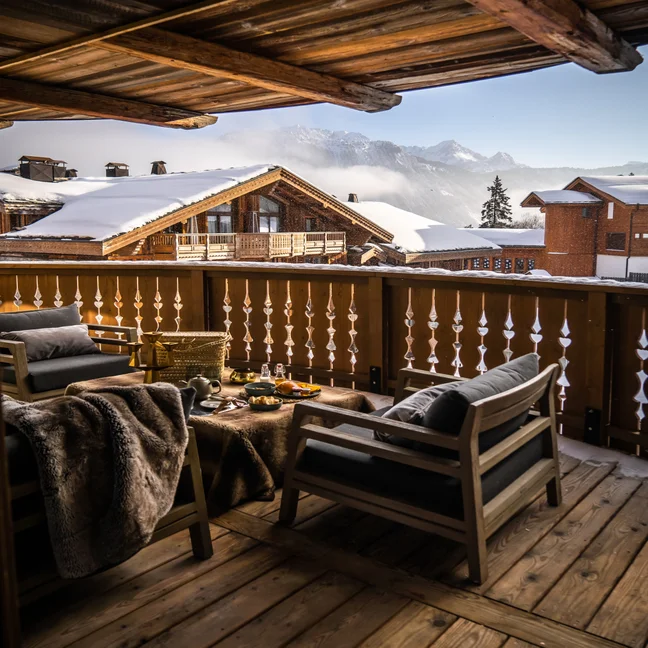 Balcon en bois avec deux fauteuils et une petite table, donnant sur les toits enneigés des chalets et les montagnes sous un ciel dégagé.