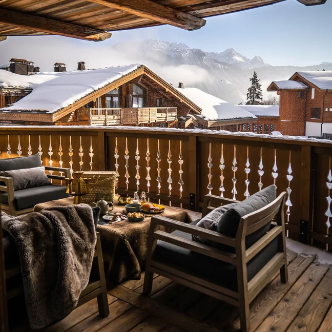 Balcon en bois avec deux fauteuils et une petite table, donnant sur les toits enneigés des chalets et les montagnes sous un ciel dégagé.