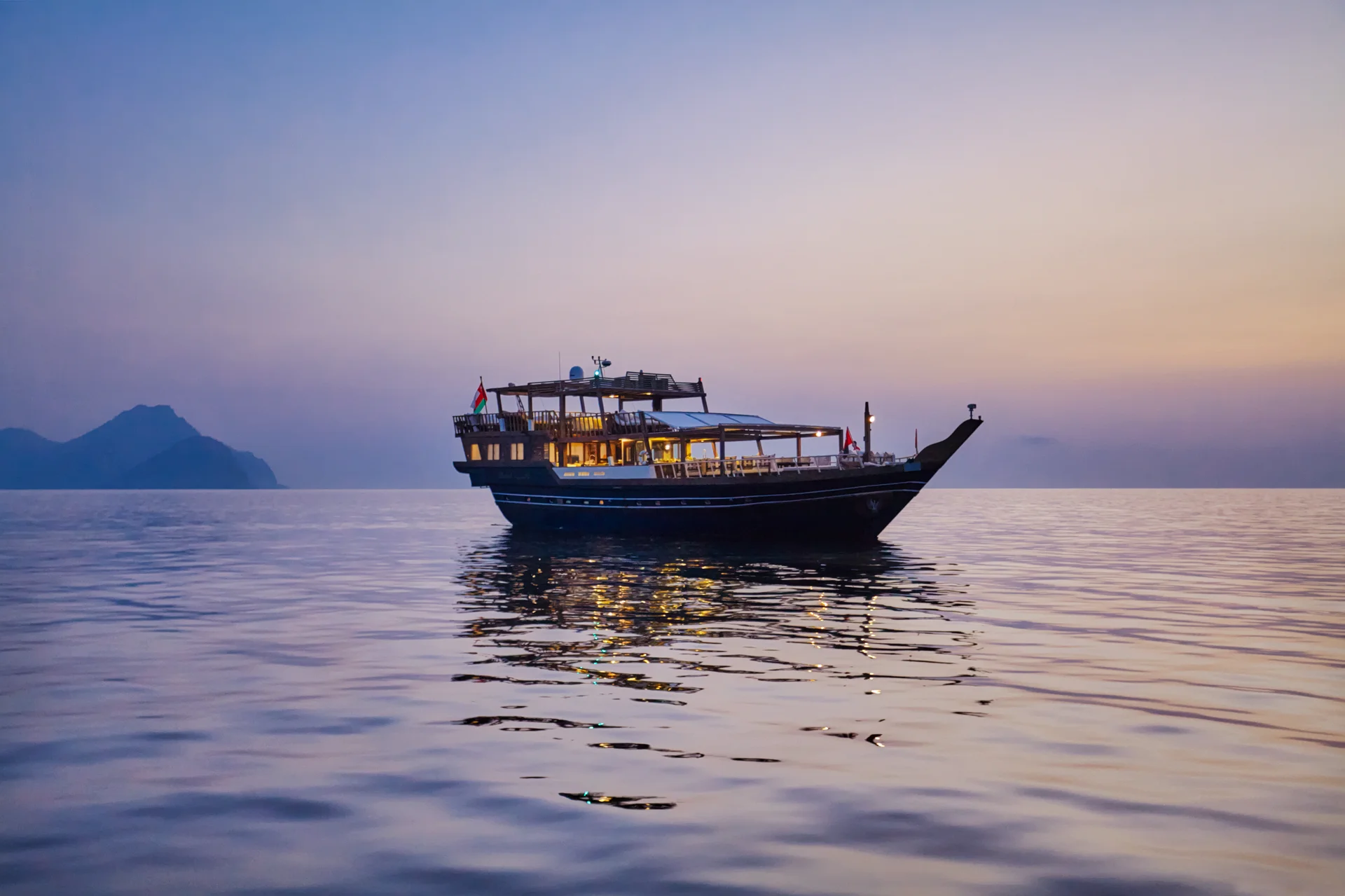 The resort's dhow Dhahab lying at anchor on flat water at first light, its lamps still lit along the deck and a headland silhouetted in the haze behind.