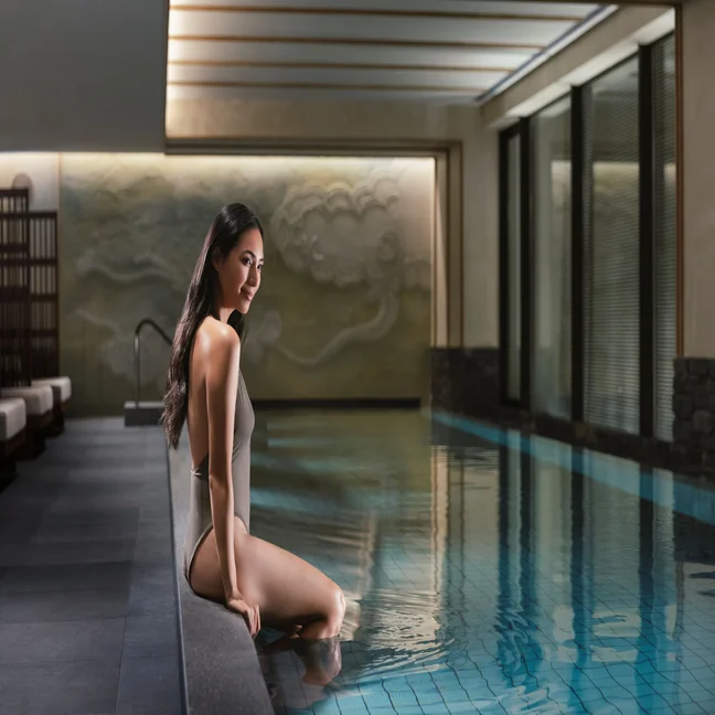 A guest relaxes at the edge of an indoor pool with legs in the water, surrounded by natural light and a tranquil spa environment designed for wellbeing.