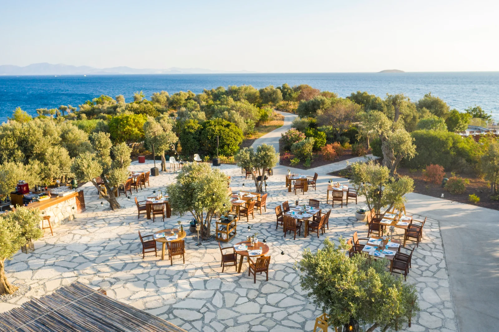 Tables laid on a stone terrace among olive trees at the water's edge, the bay curving away behind and a thatched roof in the foreground.