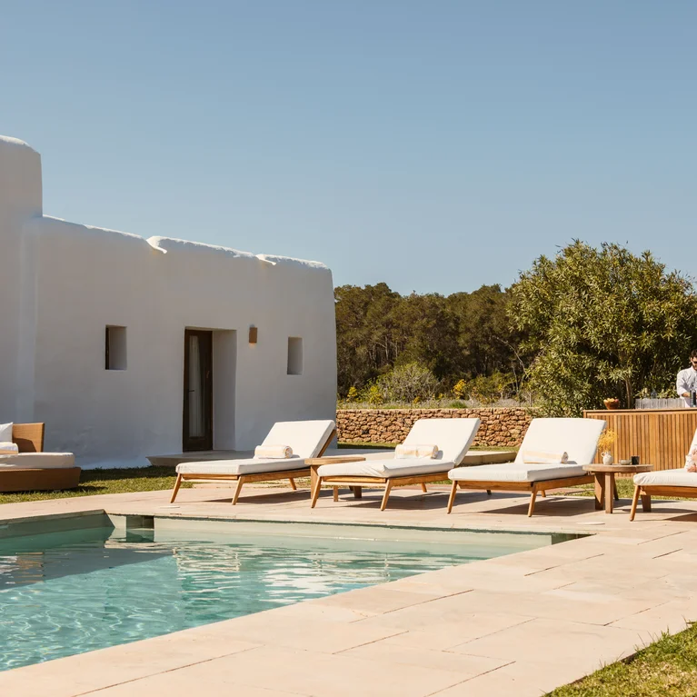 白色别墅配有游泳池、日光浴躺椅和园艺花园;在万里无云的天空下,一位客人在室外休息,另一位客人则待在树丛中的木制建筑旁。.