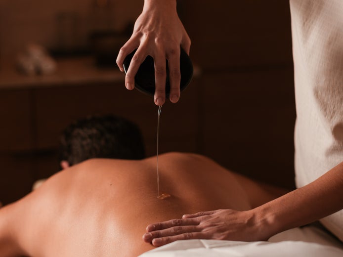 Warm oil is poured from a small bowl onto a person’s back during a spa treatment.