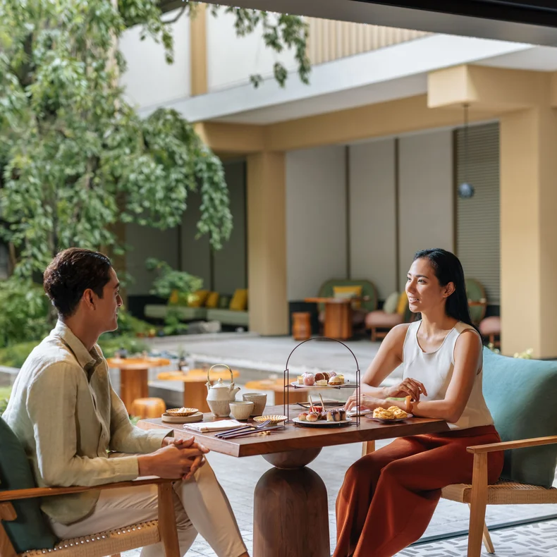 Guests share tea and pastries at an outdoor table in a tranquil garden courtyard, surrounded by comfortable seating and lush greenery.