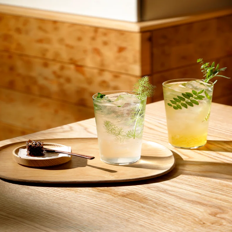 Iced beverages with fresh herbs and a chocolate cake slice on a wooden plate, arranged on a sunlit table for a balanced and refreshing experience.
