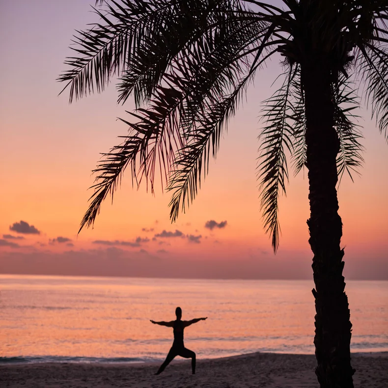 Individual doing yoga beneath a palm tree on a tranquil beach, with the ocean and sky in warm sunset tones in the background.