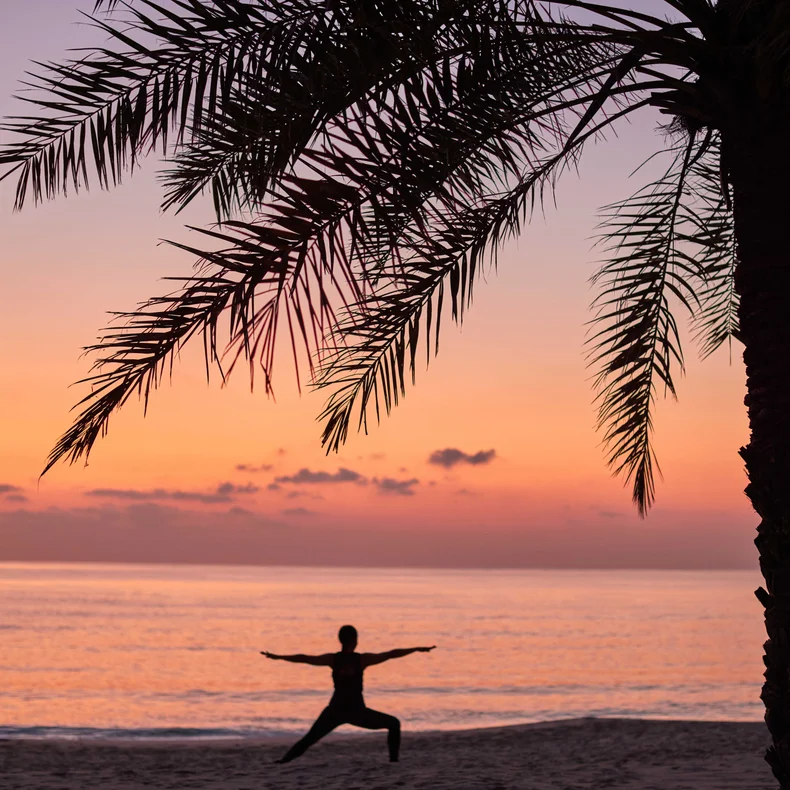 Individual doing yoga beneath a palm tree on a tranquil beach, with the ocean and sky in warm sunset tones in the background.
