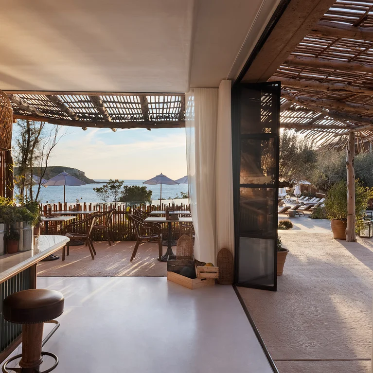 露天咖啡馆摆放着柳条桌椅,俯瞰着阳光明媚的海滩和平静的海面,木制凉棚遮阳,四周绿树环绕。.