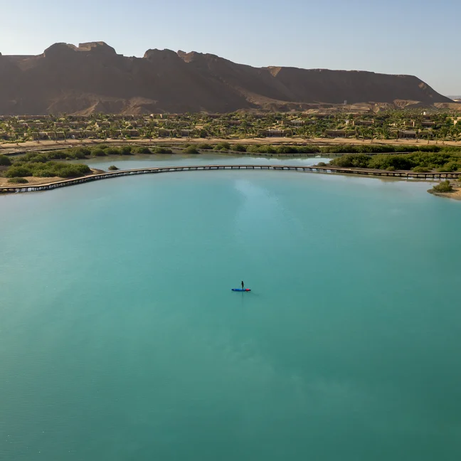 A kayaker moves across a turquoise lake bordered by green trees, with a bridge and distant mountains beneath a clear sky.
