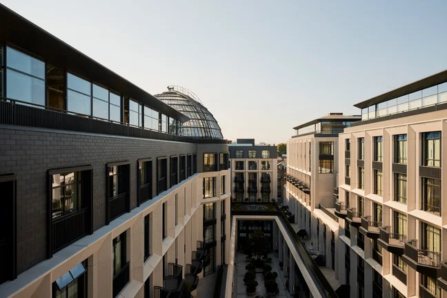 Sustainable architecture with floor-to-ceiling windows and balcony spaces surrounds a courtyard; a glass dome rises in the background under an open sky.