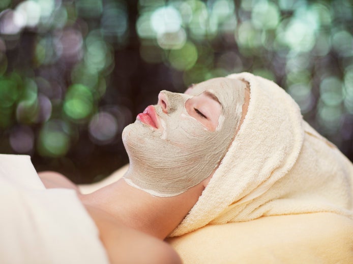 Guest with eyes closed and nourishing facial mask, hair wrapped in a towel, rests on a spa bed; soft greenery in the background enhances tranquility and focus on wellbeing.